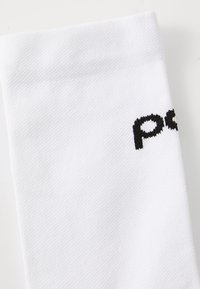 Chaussette de sport blanche en tissu texturé, avec un logo noir brodé sur le côté. Le design est élégant et de forme cylindrique.