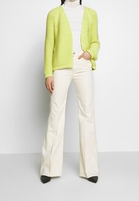 Sweter rozpinany na drutach w jaskrawym limonkowym kolorze, otwarty z przodu, o fakturowanym wzorze, z prążkowanymi mankietami i obszyciem. Połączony z kremowymi rozkloszowanymi spodniami i czarnymi szpilkami.