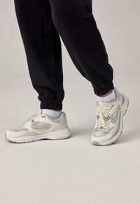 Sneakers gemaakt van beige gaas en synthetische materialen met grijze accenten, ronde neus, dikke witte zool en met patronen gedetailleerd.