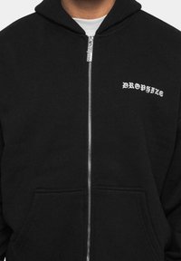 Hoodie noir zippé en mélange de coton, avec une poche frontale et le texte blanc "DROPSIZE" dans une police stylisée sur la zone du côté gauche de la poitrine.