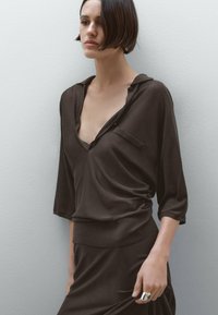Brown, lightweight blouse with a V-neck, long sleeves, and a pocket detail. Smooth texture with subtle draping effect. Model wears a matching skirt.
