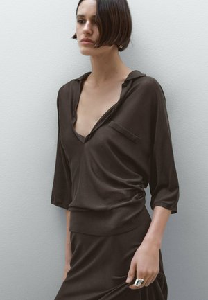 Brown, lightweight blouse with a V-neck, long sleeves, and a pocket detail. Smooth texture with subtle draping effect. Model wears a matching skirt.