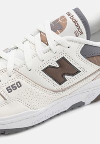 Valkoiset tennarit, joissa on rei'itetty nahka, ruskeat yksityiskohdat ja harmaat yksityiskohdat. Sisältää nauhat ja kumipohjan, jossa on "new balance" -kaiverrukset.