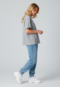 T-shirt gris à manches courtes avec une coupe ample, associé à un jeans en denim bleu clair. Le mannequin porte des baskets blanches et se tient de profil.