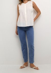 Ærmeløs hvid skjorte med knapper parret med blå slim-fit jeans og beige flade sandaler på en stående person foran en ensfarvet baggrund.