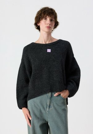 Persona con capelli corti e castani che indossa un maglione largo grigio scuro e jeans azzurri, una mano in tasca, su uno sfondo chiaro e uniforme.