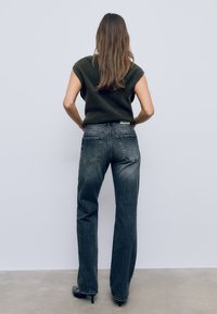 Sive kavbojske hlače s ravnim krojem, ki imajo zadnje žepe in kontrastno šivanje, v kombinaciji z temno zeleno brezrokavnico iz pletenine.