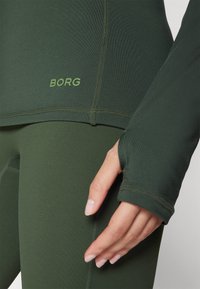 Zöld sportmárkás felső és leggings sima, testhezálló anyagból. Jellemzők közé tartozik a hüvelykujjnak való nyílás a hüvelyeken és a "BORG" logó világosabb zöld színben.