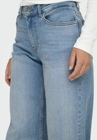 Jeans din denim de culoare bleu deschis, cu un design drept, având buzunare frontale și închidere cu un buton argintiu. Textura pare moale și ușor purtată.