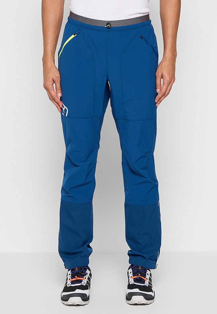 Homme portant un pantalon de randonnée bleu avec des poches zippées et des chaussures de randonnée noires et blanches, se tenant devant un fond uni.