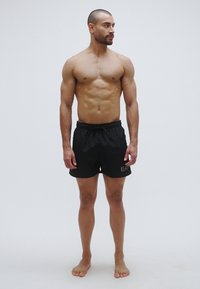 Fekete fürdőnadrág, elasztikus derékpánttal és húzózsinórral. A bal oldalon diszkrét logó látható. A modell mezítláb áll semleges háttér előtt.