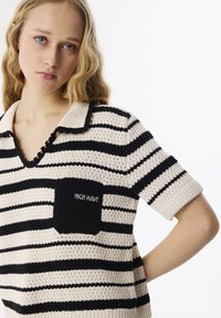 Gestreepte gebreide polo in beige en zwart, met een textuurstof, korte mouwen, gegolfde kraag en een zwarte pochet met de tekst "HIGH WAVE".