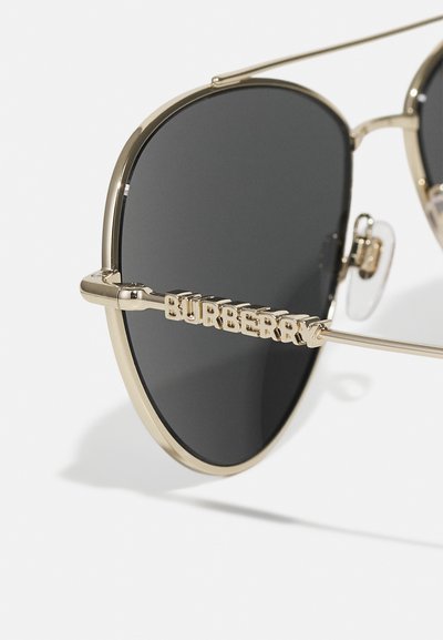 Occhiali da sole in metallo dorato con lenti nere, caratterizzati da una forma rotonda e naselli trasparenti. L'asta laterale presenta la scritta "BURBERRY" in caratteri dorati.