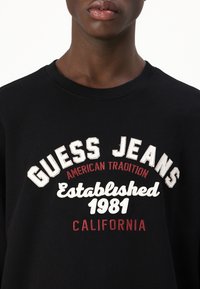 Felpa nera con testo ricamato in rilievo bianco e rosso: "GUESS JEANS," "Stabilito nel 1981" e "CALIFORNIA." Collo rotondo.