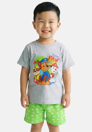 Jeune garçon souriant portant un t-shirt gris PAW Patrol et un short vert vif avec un motif blanc, debout devant un fond uni.