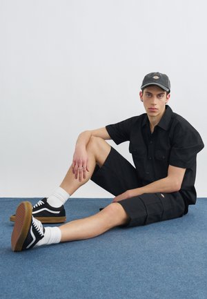Joven vestido con pantalones cortos negros, camiseta de manga corta, gorra negra, calcetines blancos y zapatillas negras, sentado sobre una alfombra azul contra un fondo liso.