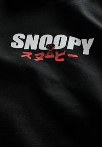 Sort stof med "SNOOPY" i fede hvide bogstaver og et rødt grafisk billede af Snoopy samt japanske tegn nedenfor.