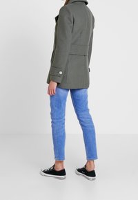 Abrigo verde oliva con un ajuste entallado, gran cuello y puños abotonados, combinado con jeans ajustados azules y zapatillas negras.