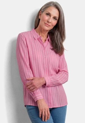 Femme aux longs cheveux gris portant une chemise rayée rose et blanche boutonnée et un jean bleu, debout, les bras légèrement croisés.