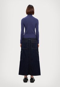 Lila gestrickter Rollkragenpullover und langer dunkler Jeansrock mit vorderen Taschen und seitlichen Schlitzen. Glatte Textur und figurbetontes Design. Chunky schwarze Schuhe.