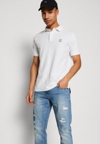 Hvid polo shirt med krave, lille logo på brystet, parret med lyseblå ripped jeans og en sort cap. Glat stof tekstur.