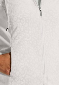 Witte hoodie met ritssluiting, voorzien van een gestructureerd luipaardprintpatroon. De stof lijkt zacht en pluche, met zilverkleurige hardware en trekkoordaccenten.