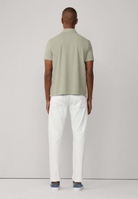 Polo a maniche corte di colore verde chiaro, abbinato a jeans bianchi. Vista posteriore che mostra il colletto della maglietta e la vestibilità rilassata, indossata con scarpe da ginnastica blu.