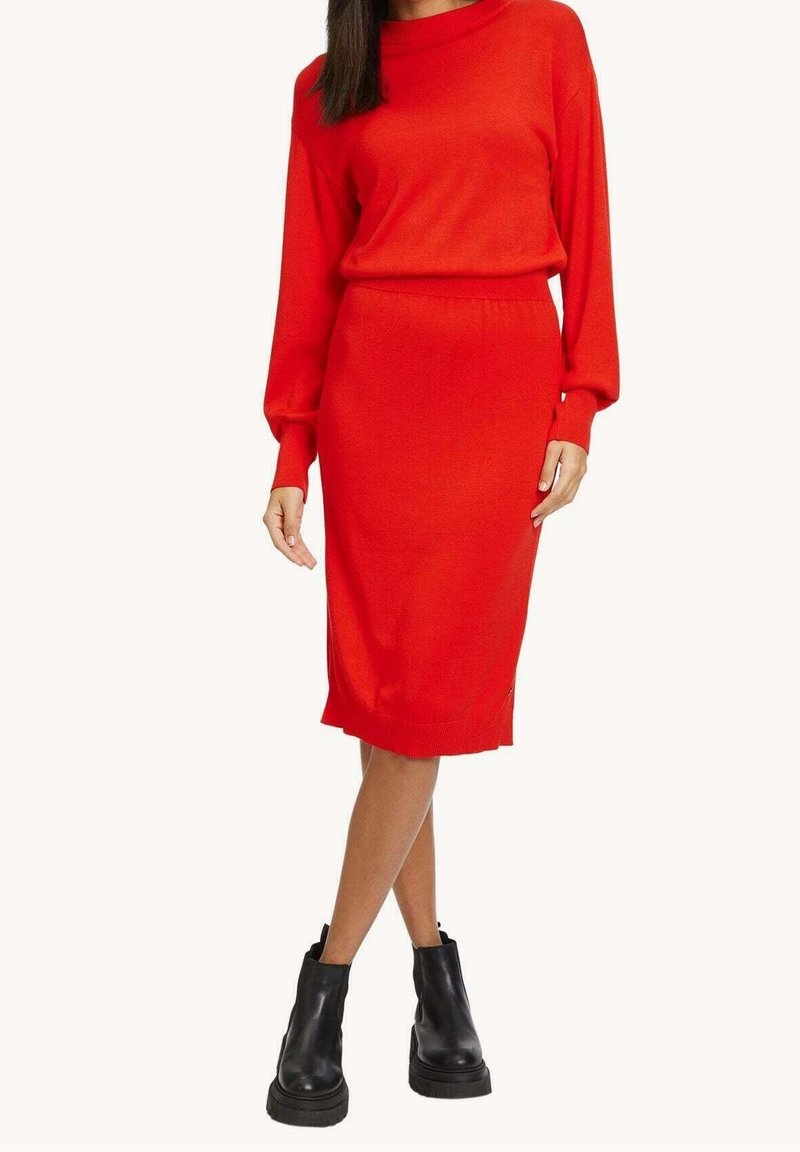 Femme portant une robe en maille rouge à manches longues associée à des bottines noires épaisses, debout devant un fond uni.