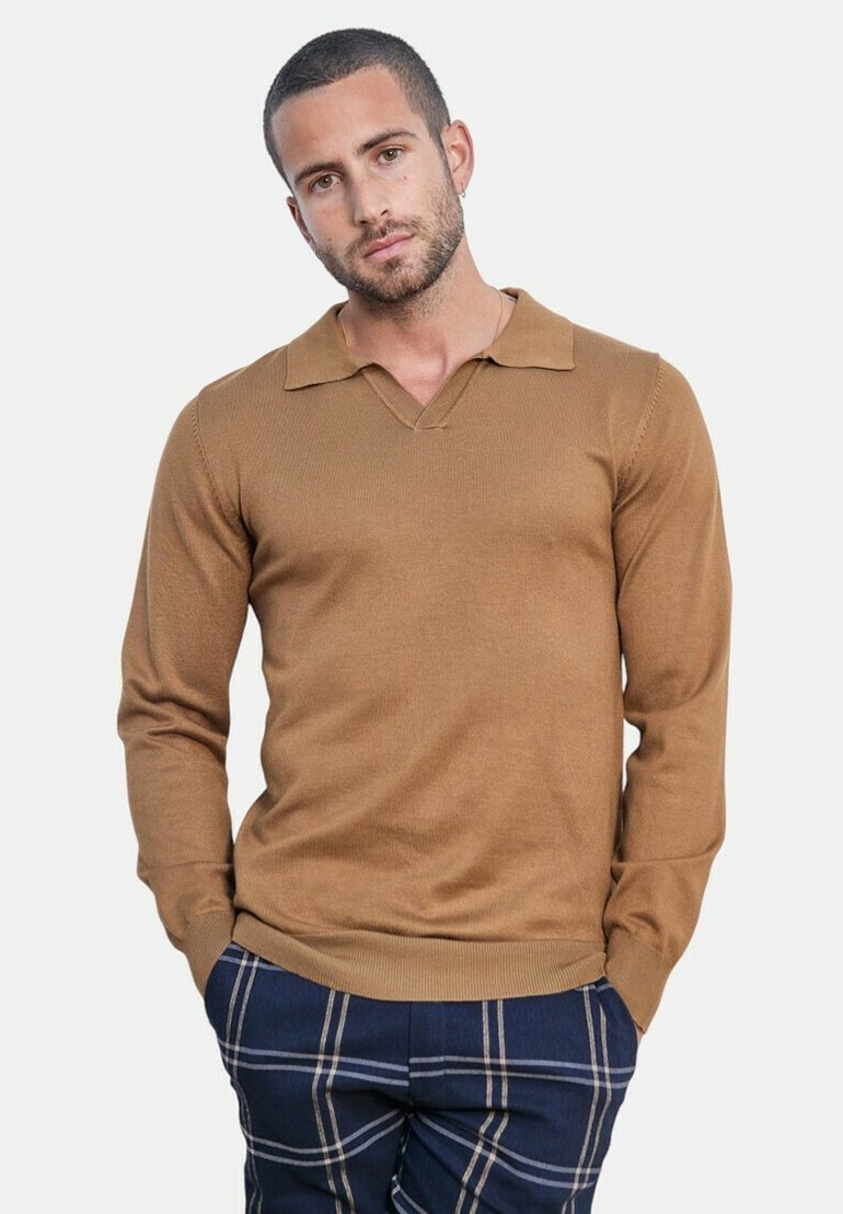 Homme aux cheveux courts et à la barbe, portant une chemise marron à manches longues avec col et un pantalon à carreaux bleu, debout les mains dans les poches.