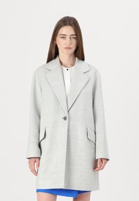 Cappotto in lana grigio chiaro con colletto a revers, chiusura con un solo bottone e due tasche frontali. Le maniche sono leggermente oversize.
