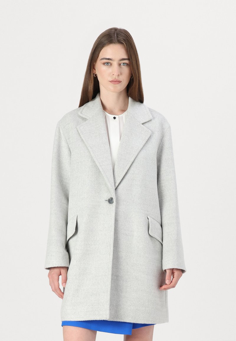Cappotto in lana grigio chiaro con colletto a revers, chiusura con un solo bottone e due tasche frontali. Le maniche sono leggermente oversize.