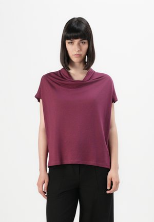 Donna con capelli scuri e lisci indossa una maglietta corta e ampia di colore bordeaux con scollo drappeggiato e pantaloni neri su uno sfondo bianco.