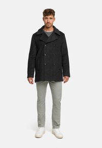 Abrigo de lana negro tipo peacoat con diseño de doble botonadura, solapas anchas y dos bolsillos laterales, combinado con pantalones gris claro y zapatillas blancas.
