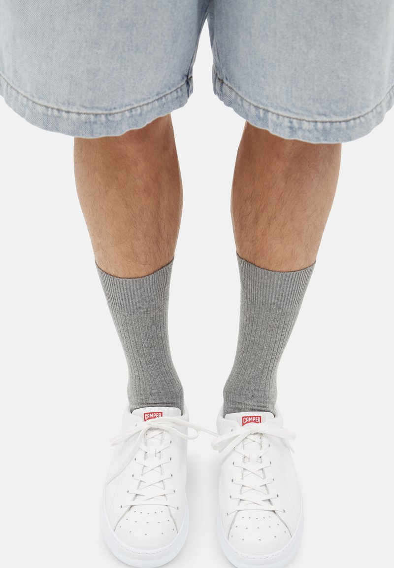 Sneaker bianchi in pelle, calzini grigi a coste e pantaloni shorts in denim azzurro. Le sneakers presentano un accento rosso del logo e un dettaglio perforato sulla punta.