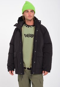 Zwarte pufferjas met een capuchon, voorzien van drukknoopsluitingen en zijzakken. Draag het over een groene shirt met een zwart grafisch ontwerp, gecombineerd met olijfgroene broek.