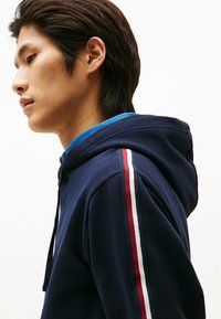 Granatowy hoodie ze sznurkiem, z niebieską koszulą pod spodem oraz paskami w kolorach czerwonym, białym i zielonym na rękawach. Miękka faktura.