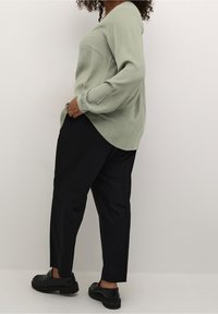 Langärmlige, hellgrüne Bluse mit strukturiertem Finish und V-Ausschnitt, kombiniert mit geraden schwarzen Hosen und klobigen schwarzen Schuhen.