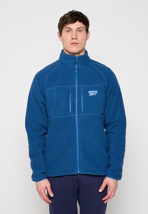 Uomo che indossa una giacca in pile blu con zip e logo Reebok, in piedi davanti a uno sfondo chiaro uniforme, rivolto in avanti.