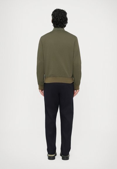 Oliviroheline jakk soonikkoesäärte ja -varrukatega, millel on seismiskaelus. Ühendatud tumesiniste pükste ja mustade kingadega. Vaade tagant.