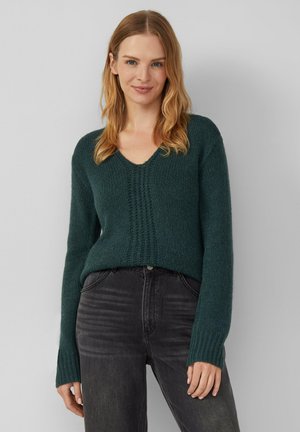 Waldgrüner Strickpullover mit V-Ausschnitt und Mittelnahtdetail, langen gerippten Ärmeln, kombiniert mit schwarzen hoch taillierten Jeans.