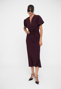 Burgundi midi suknelė su giliu V formos iškirpimu, trumpomis pūstomis rankovėmis, suktais priekiniais detalėmis, prigludusia siluetu ir švelnia medžiaga. Derinama su juodais aukštakulniais.