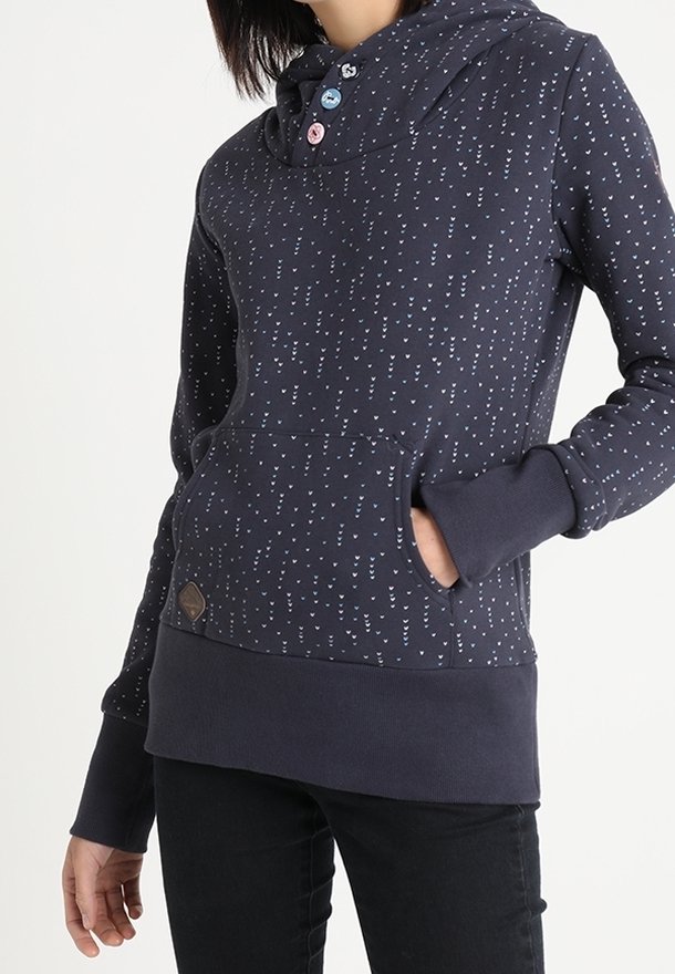 Femme portant un sweat à capuche bleu marine avec de petits motifs de flèches blanches, trois boutons près du col, poche avant et pantalon noir.