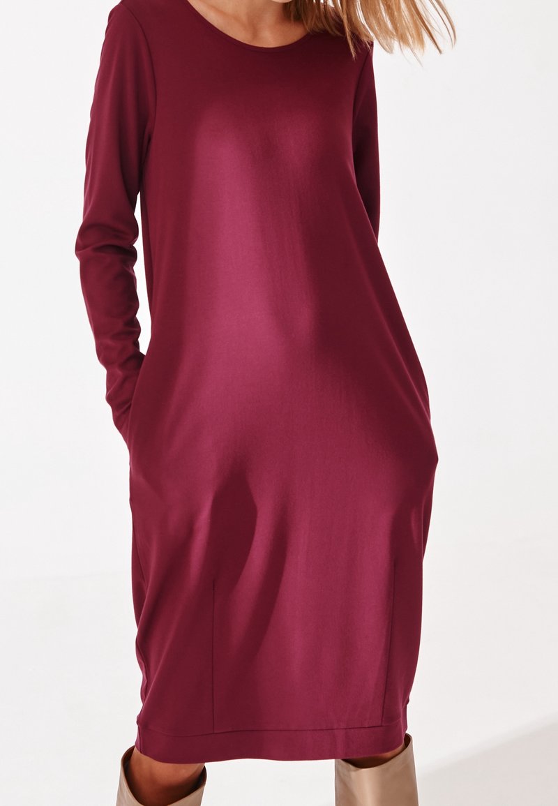 Bordowa sukienka z długim rękawem i okrągłym dekoltem, z bocznymi kieszeniami i luźnym krojem, który opada nieco poniżej kolana. Gładka tekstura.