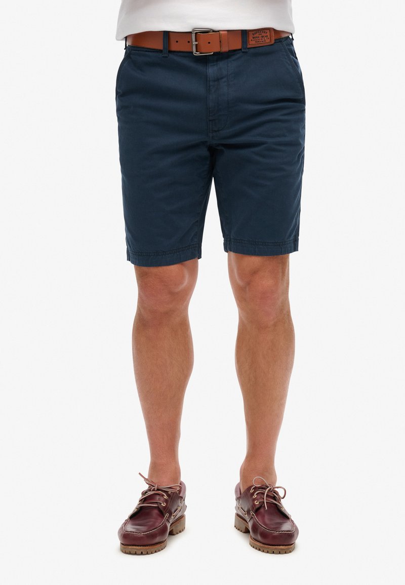 Navyblauwe katoenen shorts met een vlakke voorkant, zijzakken en een bruine leren riem. Bruine boot schoenen met veters maken de outfit compleet.