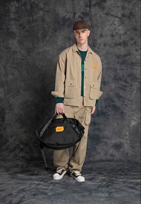 Completo beige a due pezzi con giacca e pantaloni, caratterizzato da un piccolo etichetta arancione. Borsa a tracolla nera con tracolle regolabili e accenti arancioni. Cappello marrone.