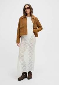 Bruine suède cropped jack over een witte kanten maxi-jurk. Donkerbruine enkellaarzen met zilveren accenten. Model draagt een zonnebril.