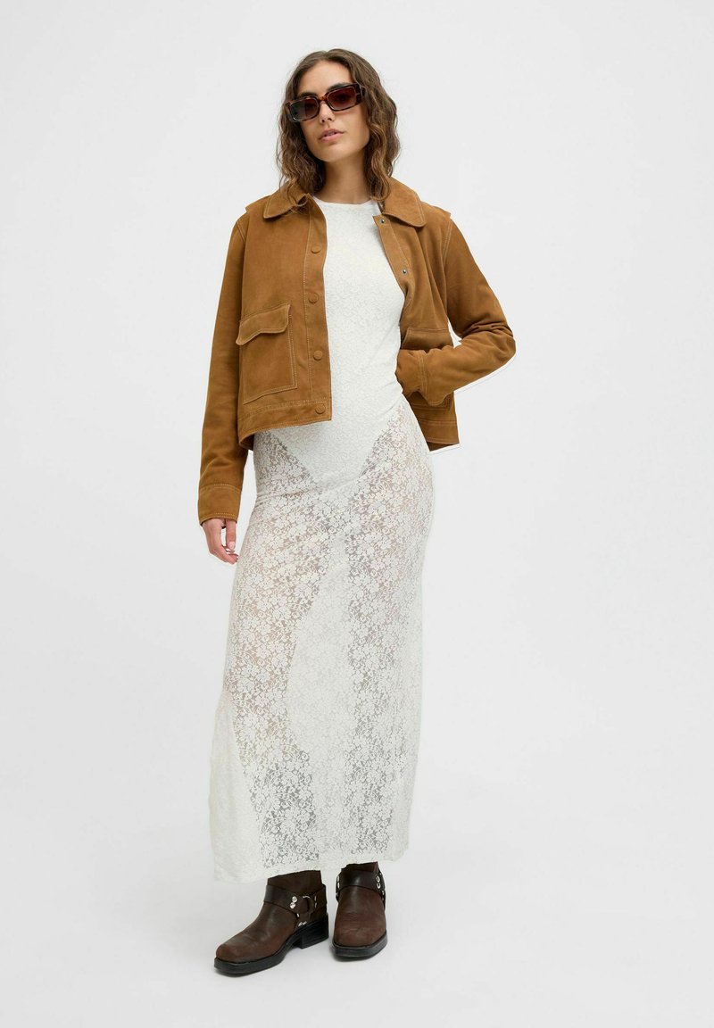 Bruine suède cropped jack over een witte kanten maxi-jurk. Donkerbruine enkellaarzen met zilveren accenten. Model draagt een zonnebril.