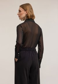 Sort gennemsigtig langærmet skjorte med krave, parret med sorte højlivet bukser med elastisk talje og sidelommer.