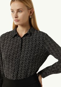 Schwarzes Button-up-Hemd mit einem geometrischen Goldmuster, spitzem Kragen und langen Ärmeln. Glattes Material mit taillierter Passform.