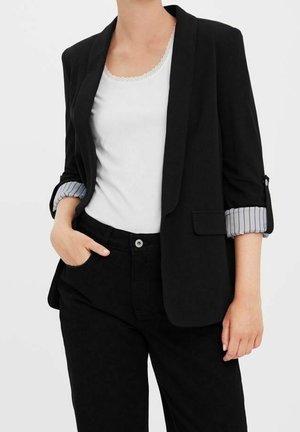 Personne portant un blazer noir avec des manches rayées retroussées, un débardeur blanc orné de dentelle, et un pantalon noir avec une main dans la poche.